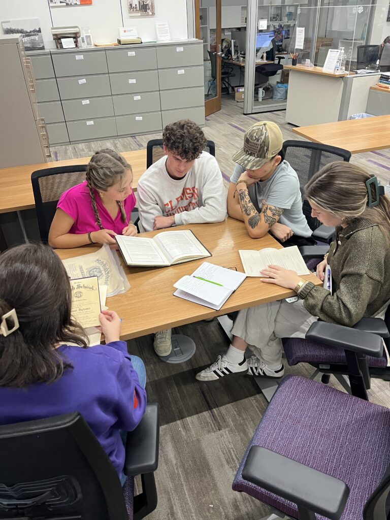 A group of students sit around the end of a table looking at notebooks.