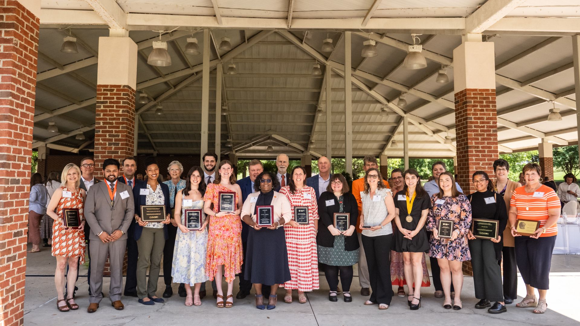 A large group of people stand under the bleachers of Memorial Stadium with the ones in the front row holding plaques.