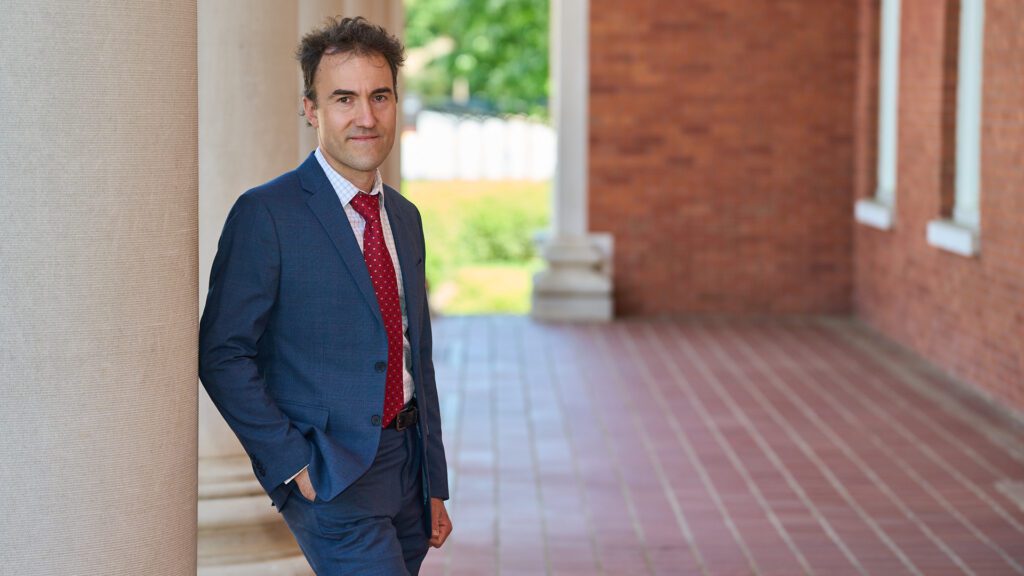 Man wearing suit leans up against a column in front of Sikes Hall at Clemson.