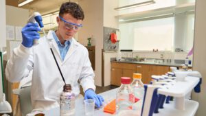 man wearing goggles and a blue and white shirt with a white lab coat pipettes in a science lab
