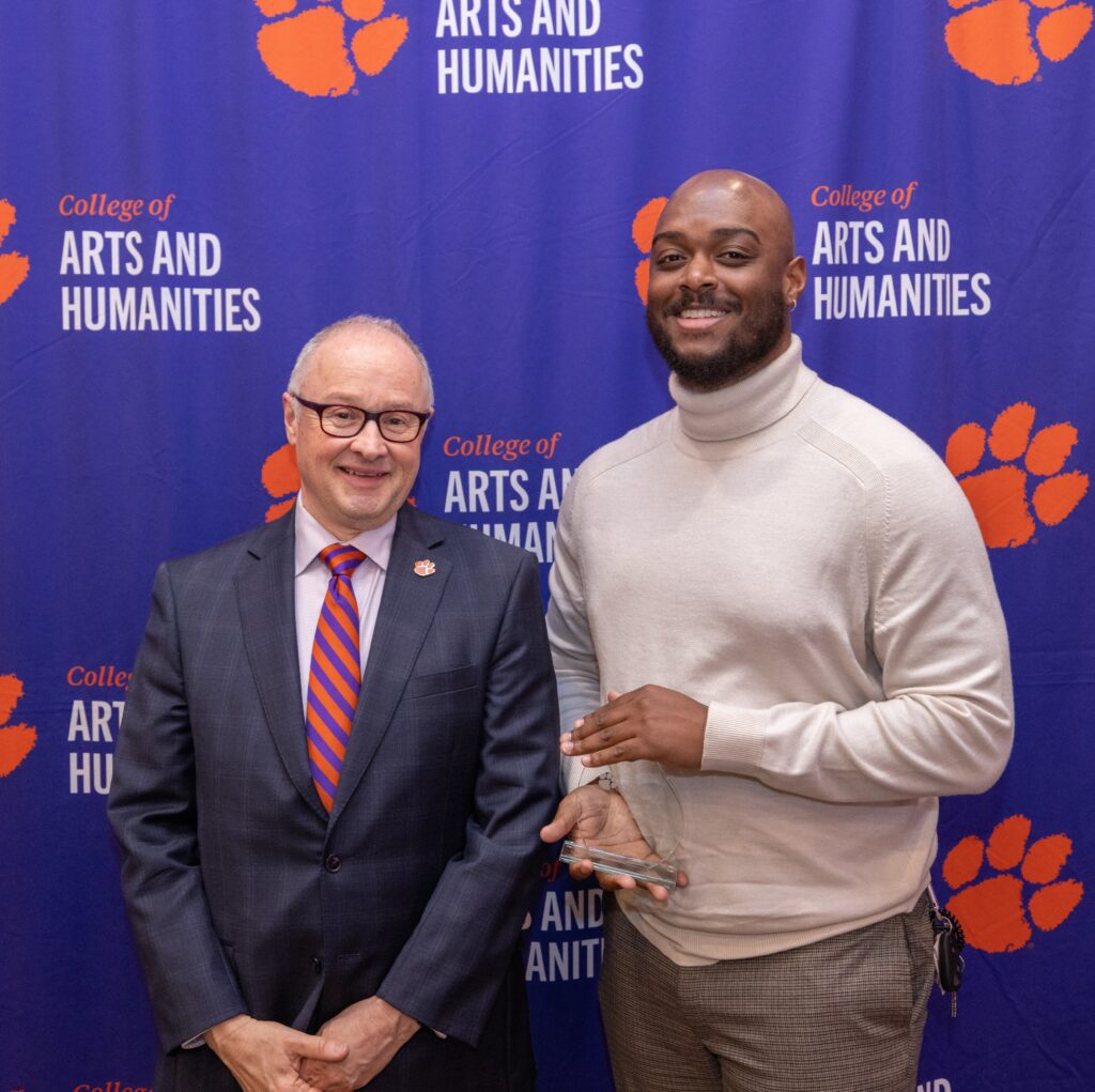Two men stand in front of a College of Arts and Humanities banner and smile at the camera, the man on the left holding a glass award.