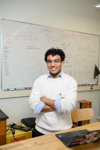 Fermin Redondo is wearing a white sweater with blue cuffs rolled up at the sleeves and he poses for a photo with arms crossed in front of a whiteboard