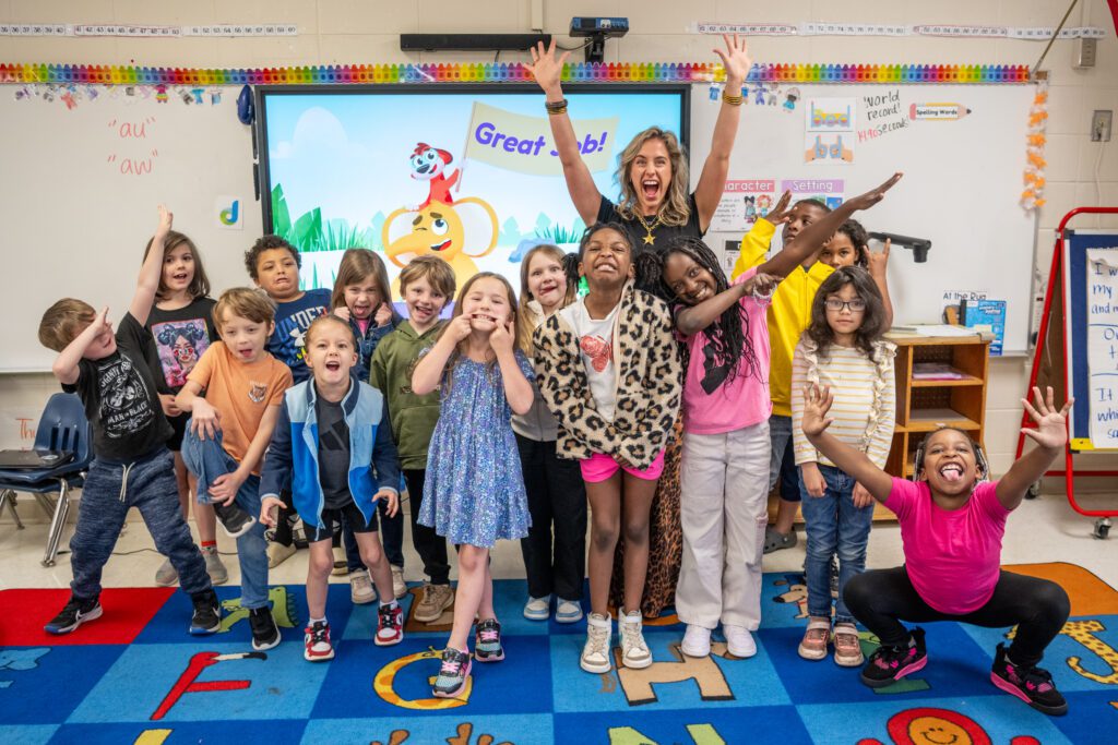 A group of elementary school kids make funny faces at the camera while their teacher raises her arms in the air and laughs behind them