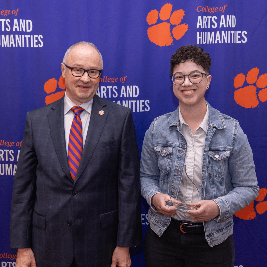 Two people stand in front of a College of Arts and Humanities banner and smile at the camera, the person on the left holding a glass award.