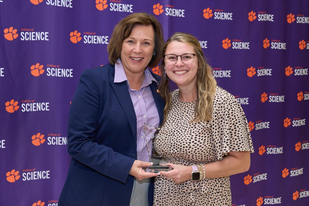 Two women stand in front of a Clemson College of Science step and repeat banner holding a clear acrylic plaque