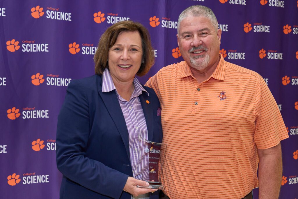 A woman and a man stand in front of a Clemson College of Science banner holding a clear acrylic trophy.