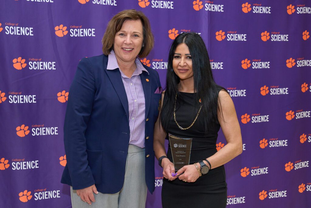 Two women stand in front of a College of Science banner and one holds a plaque.