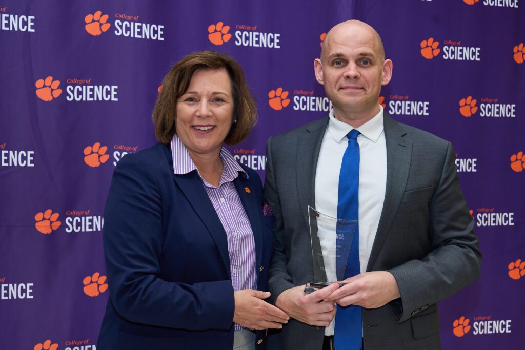 A woman and a man stand in front of a Collegeof Science banner holding a trophy