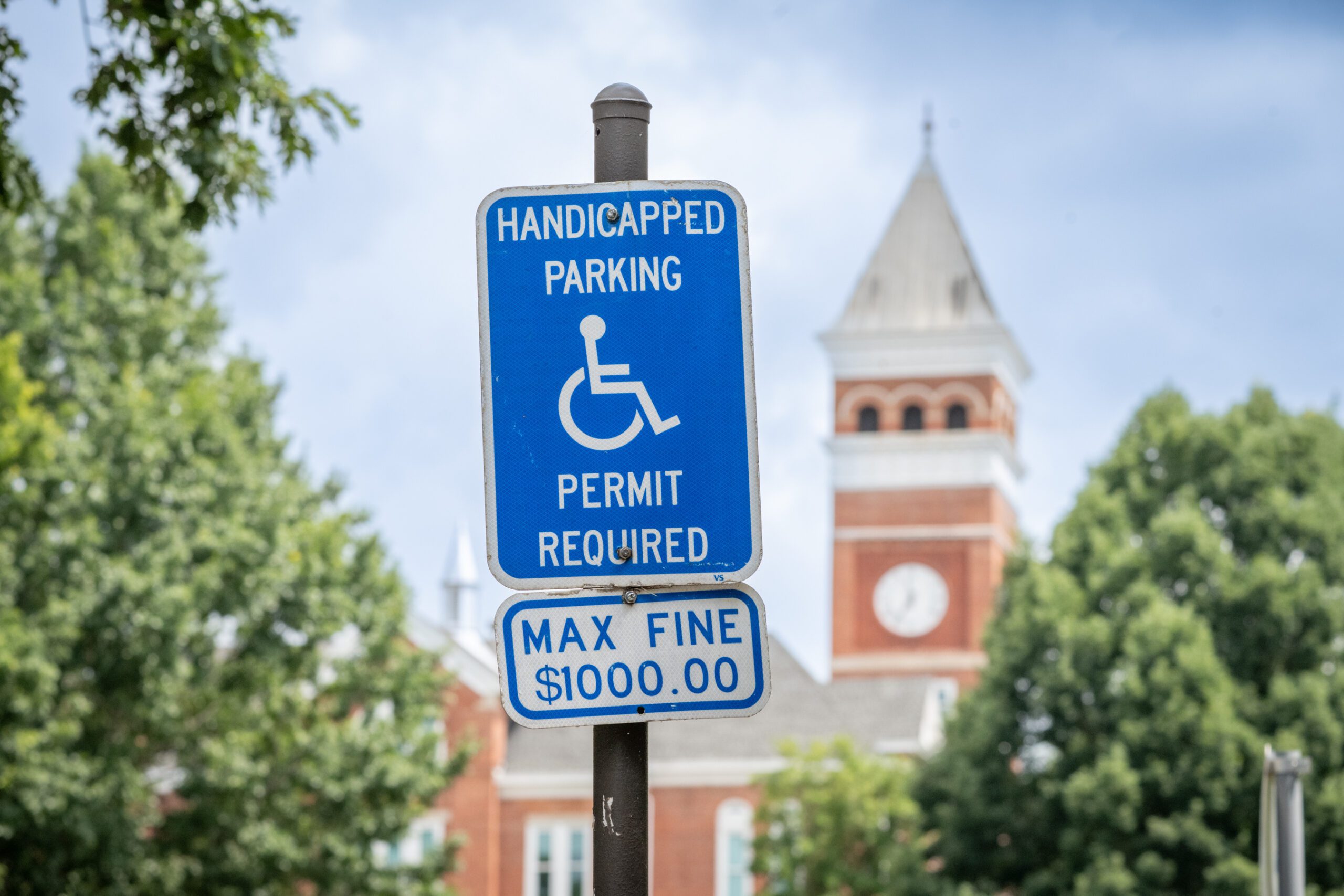 A handicapped parking sign against a blue sky with the bell tower of Tillman Hall visible behind it