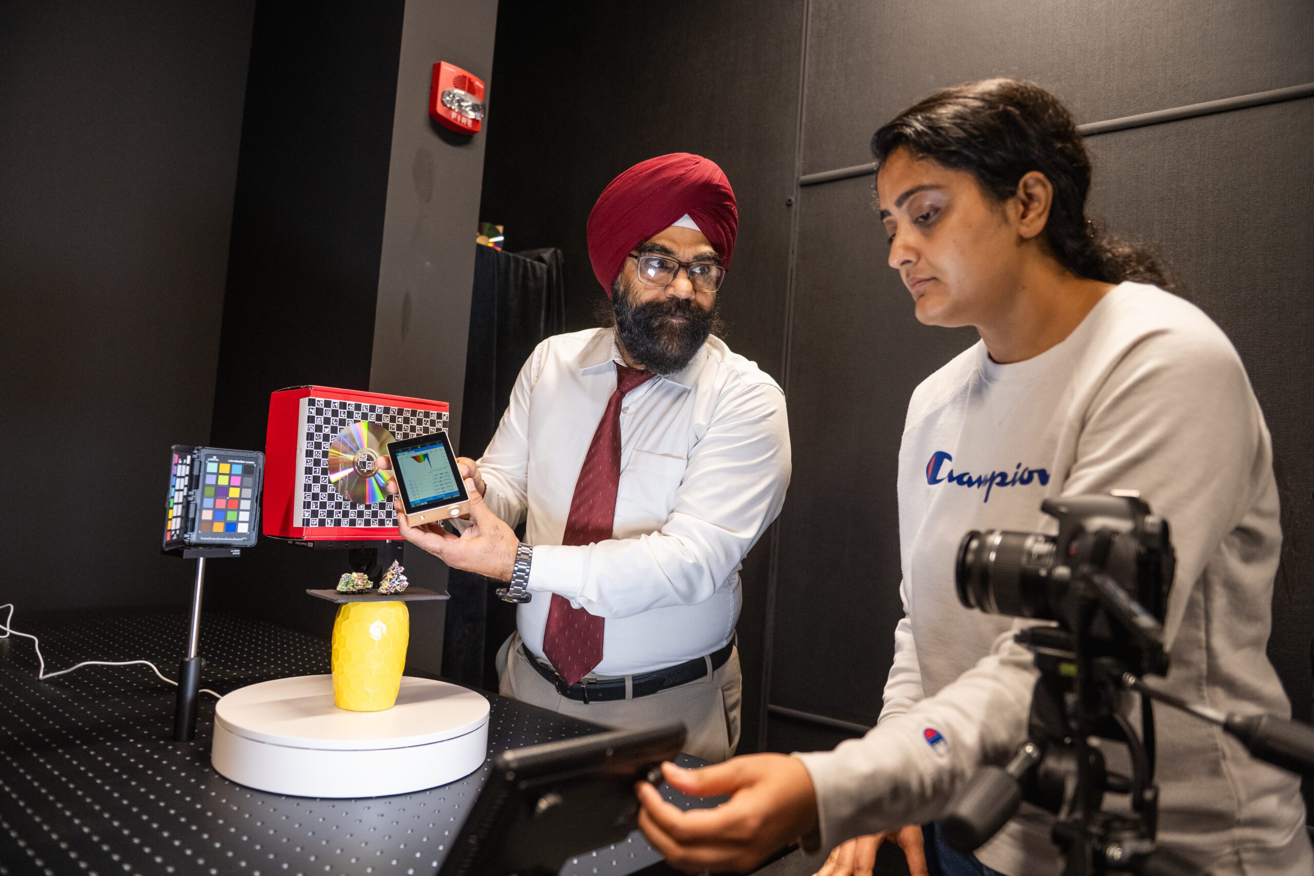 A bearded man in a turban and a young woman work together photographing small objects in a studio.