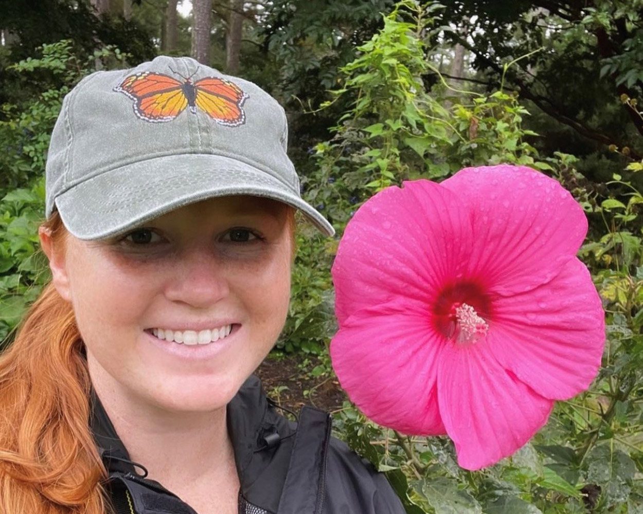 A woman with long red hair wearing a baseball cap with a butterfly on it stands next to a large pink flower that's as big as her head.
