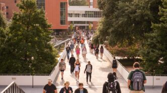 A large group of college students, many carrying backpacks, walks across the Library Bridge on the Clemson University campus. In the background, an academic building with glass and brick exterior walls is visible.