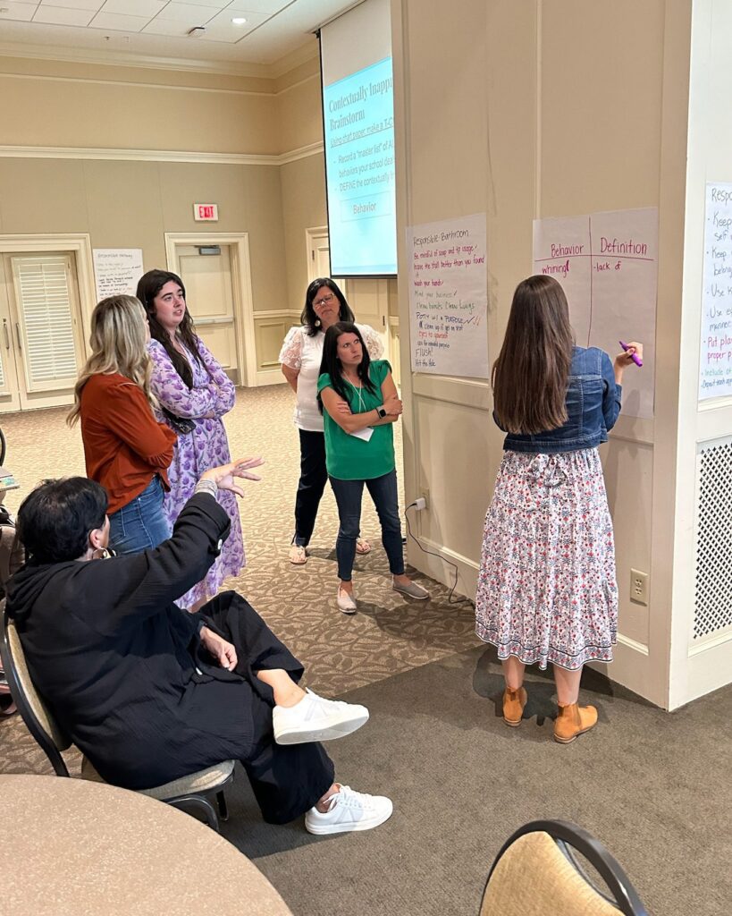 Educators stand around a large piece of white paper that they are filling out with two columns representing "behavior" and "definition."