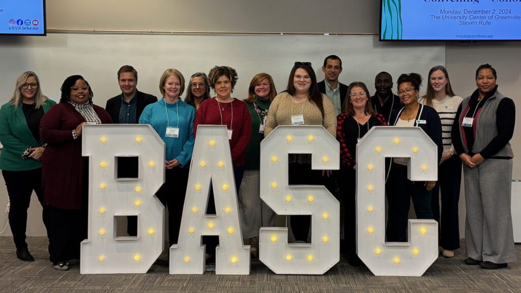A group of educators stands behind a large block of letters spelling out "BASC."