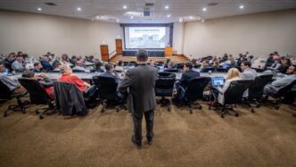 A man in a dark suite stands with his back to the camera in the back of an auditorium full of people looking at a screen in the front of the room that has the words "Mass Timber Hospital" and a rendering of a hospital on it.