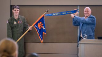 Two man stand next to each other, one holding a pole with a guidon on it and the other holding a streamer attached to the pole