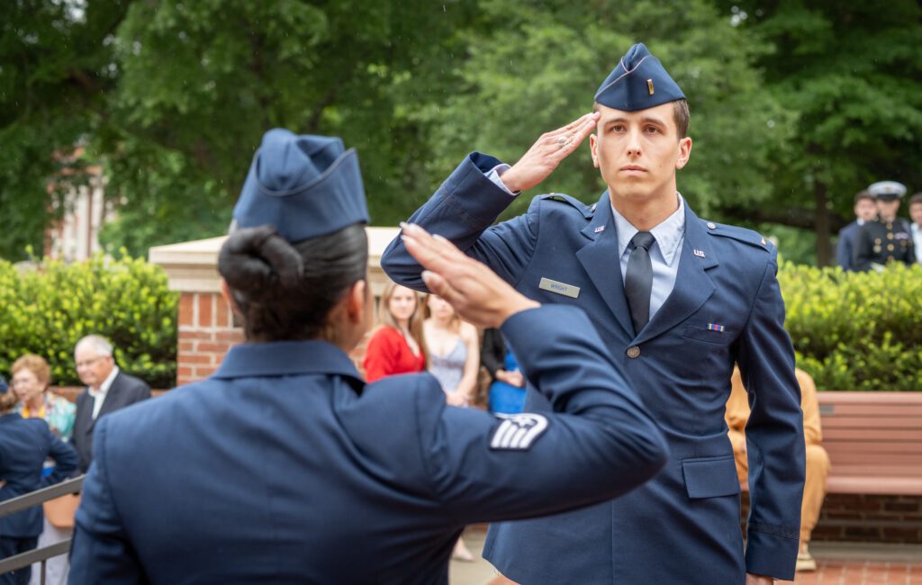 Two people dressed in formal Air Force dress uniforms salute each other, one standing with their back to the camera and the other facing the camera.