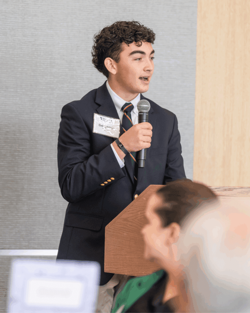 A young man in a navy blazer and tie speaks at a podium