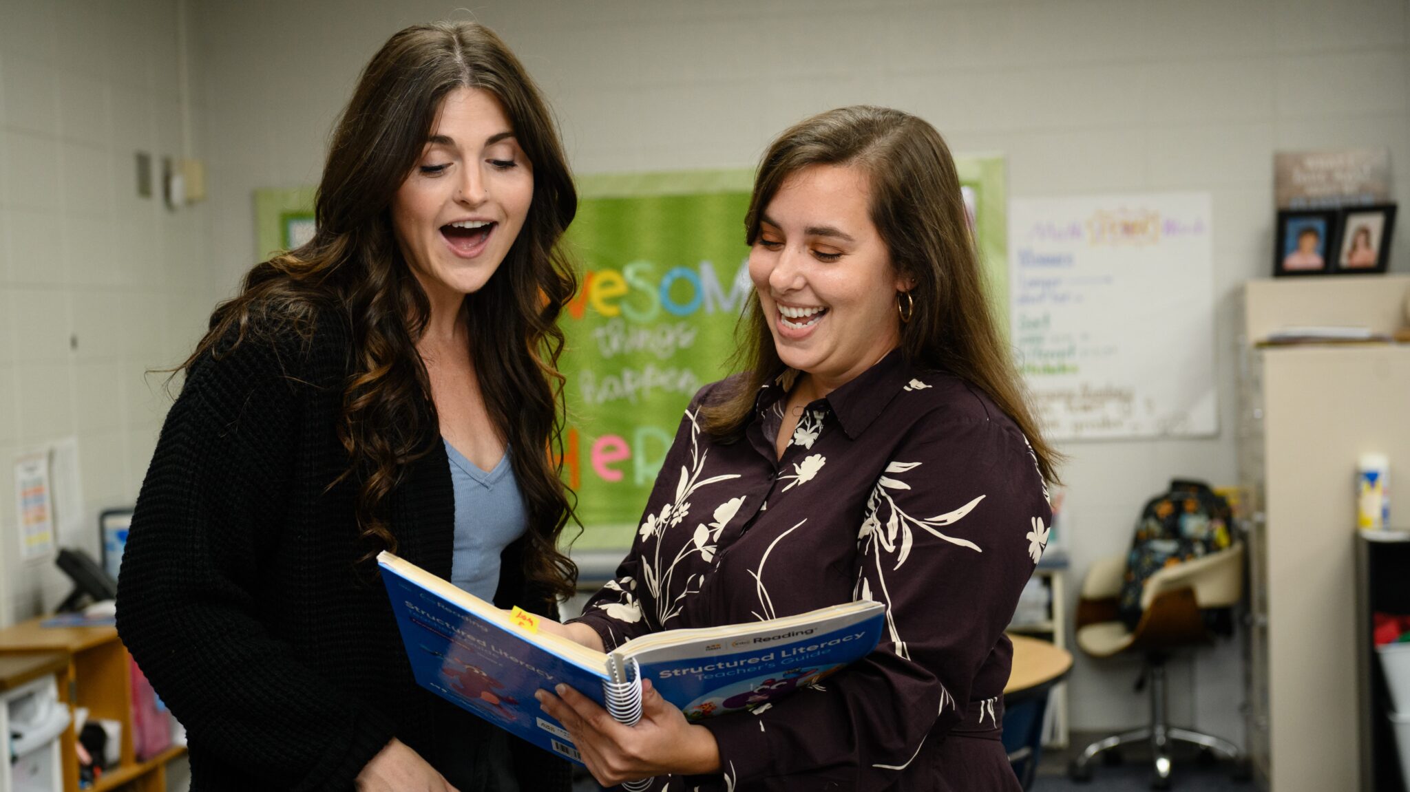 Two women, one younger the other older, both with long dark hair are in an elementary school classroom and they are laughing and looking at a spiral bound textbook.