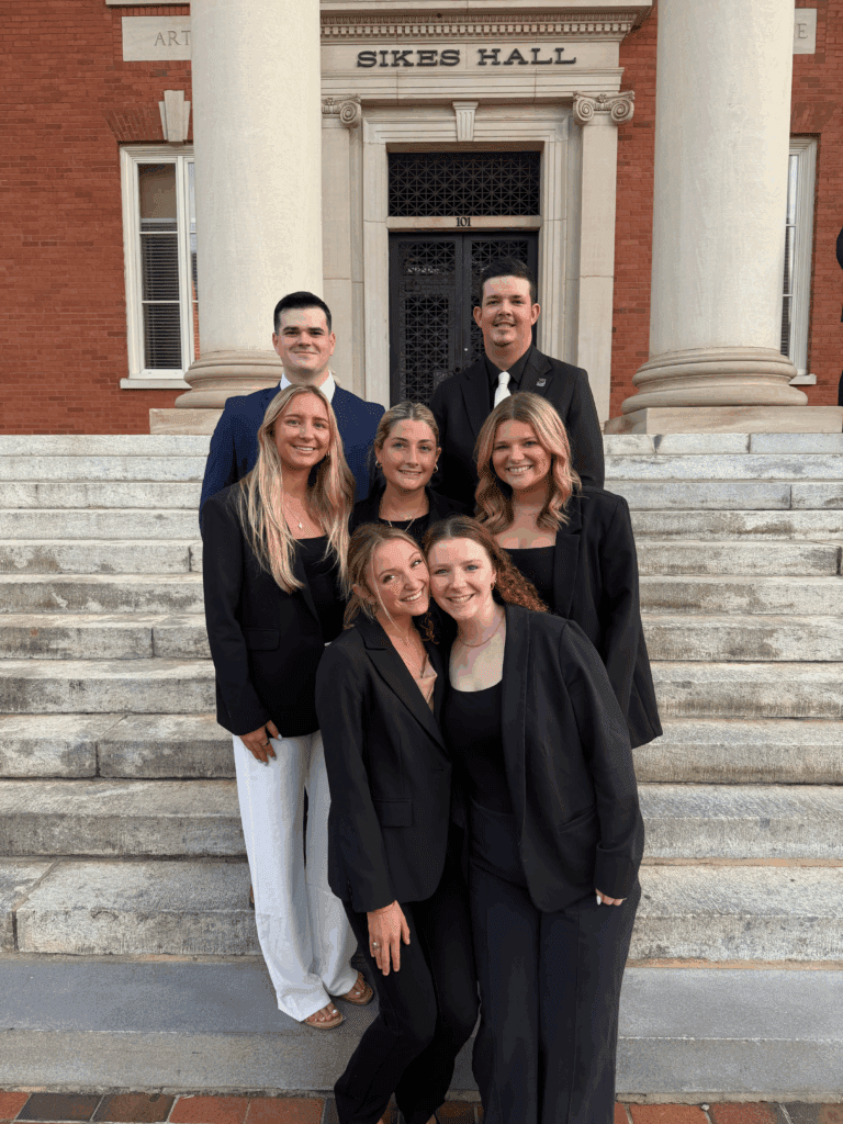 Students in dark business attire stand on the steps of Sikes Hall