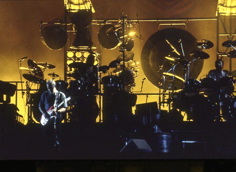 A guitarist stands in the spotlight playing while golden lights illuminate the band behind him.