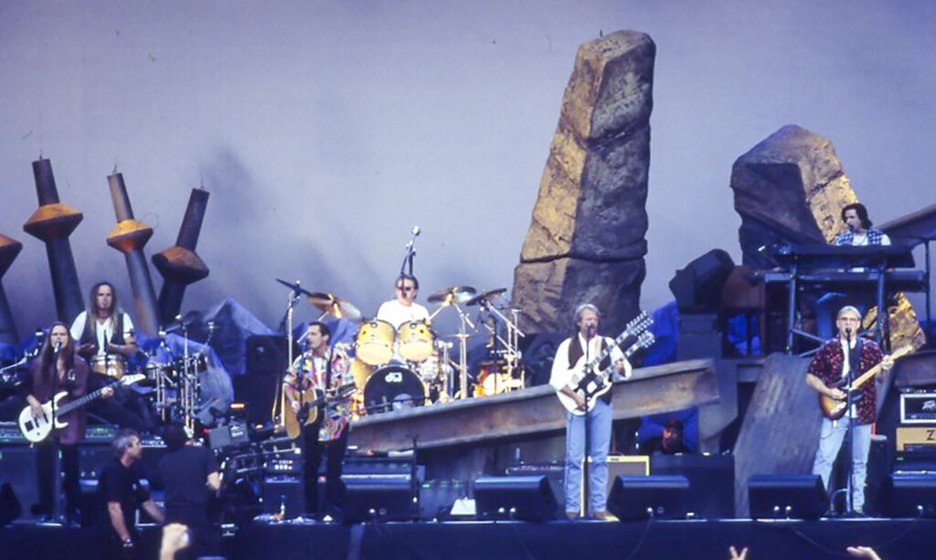 A band plays in front of a set that looks like rock formation and large metal vehicle axles standing upright.