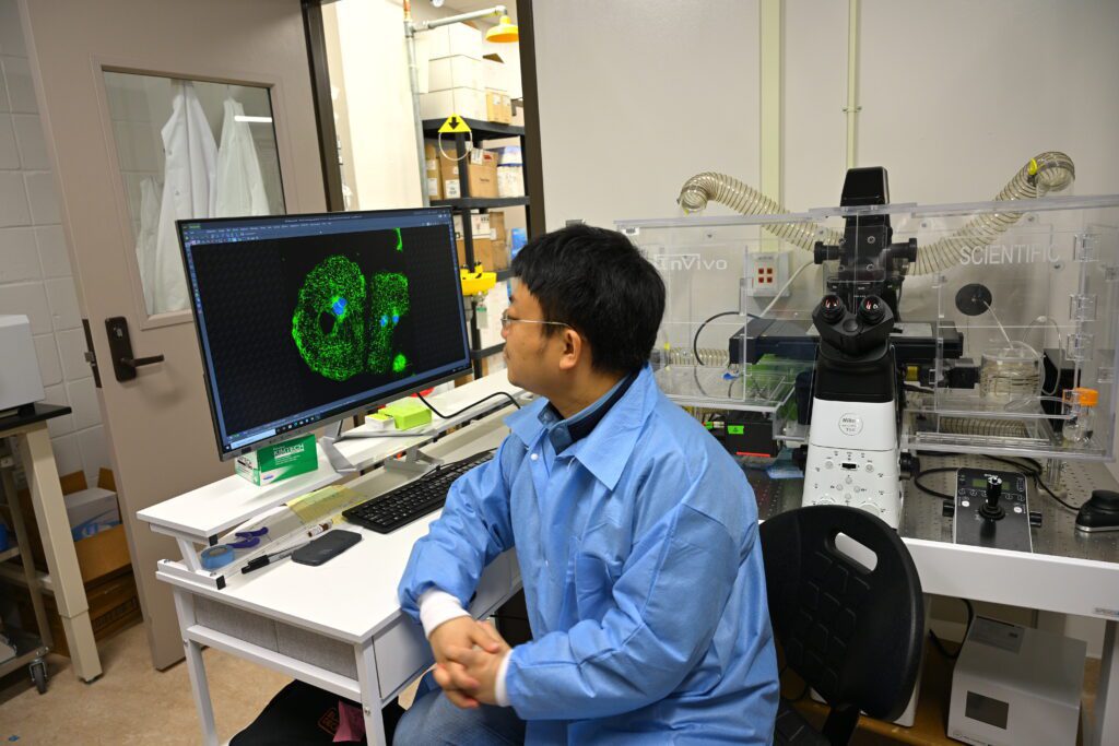 man wearing blue lab coat sits at desk looking at a green and blue diagram on his monitor